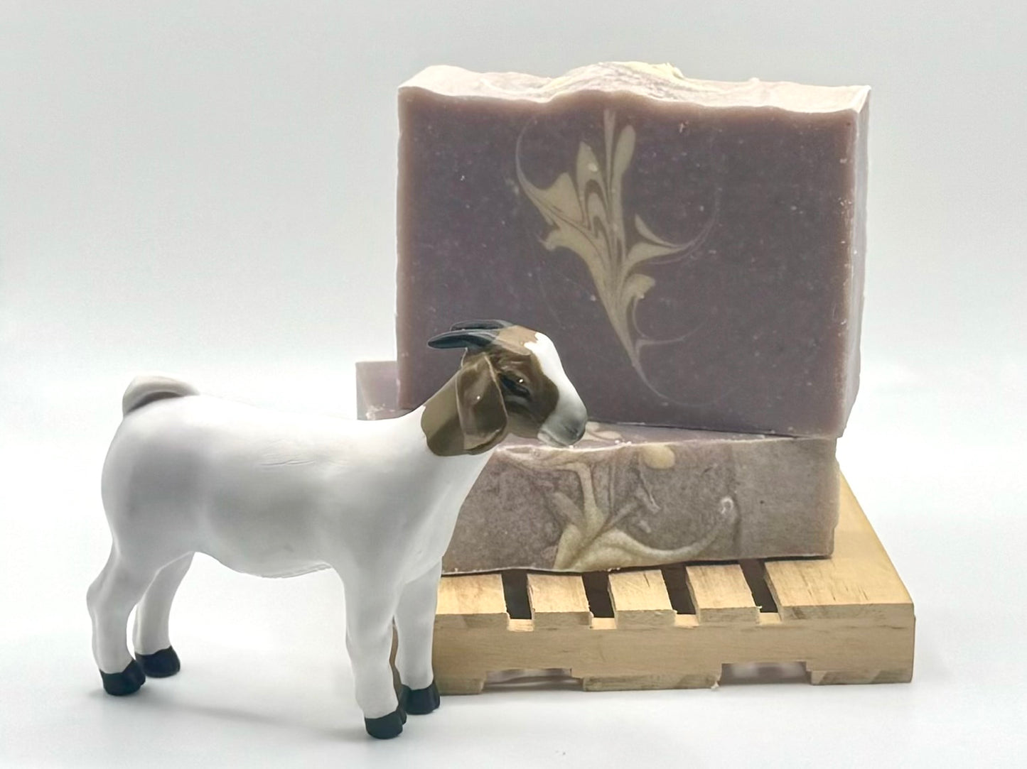 Two purple bars of soap with a white swirl are resting on a wooden block. The top bar is facing the viewer. The bottom bar only has the top visible. A goat is in the foreground.