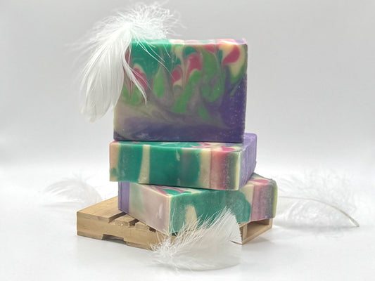 Three stacked handmade soap bars with a marbled swirl pattern of green, purple, pink, and cream, decorated with white feathers and displayed on a small wooden stand against a white background.