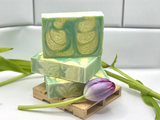 Three stacked rectangular bars of handmade soap in light green and white marbled swirls, displayed on a small wooden block with a purple tulip flower resting in front, against a white tiled background.