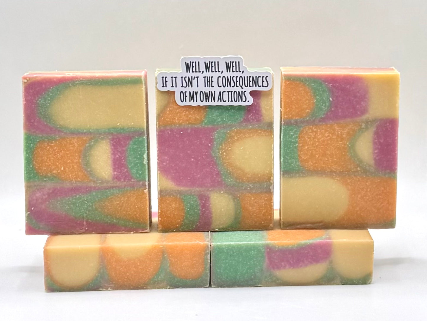 Five handmade rectangular bars of soap with a marbled swirl pattern of orange, purple, green, and cream, arranged on a white background. The center bar has a small white paper label with black text that reads, 'WELL, WELL, WELL, IF IT ISN'T THE CONSEQUENCES OF MY OWN ACTIONS.