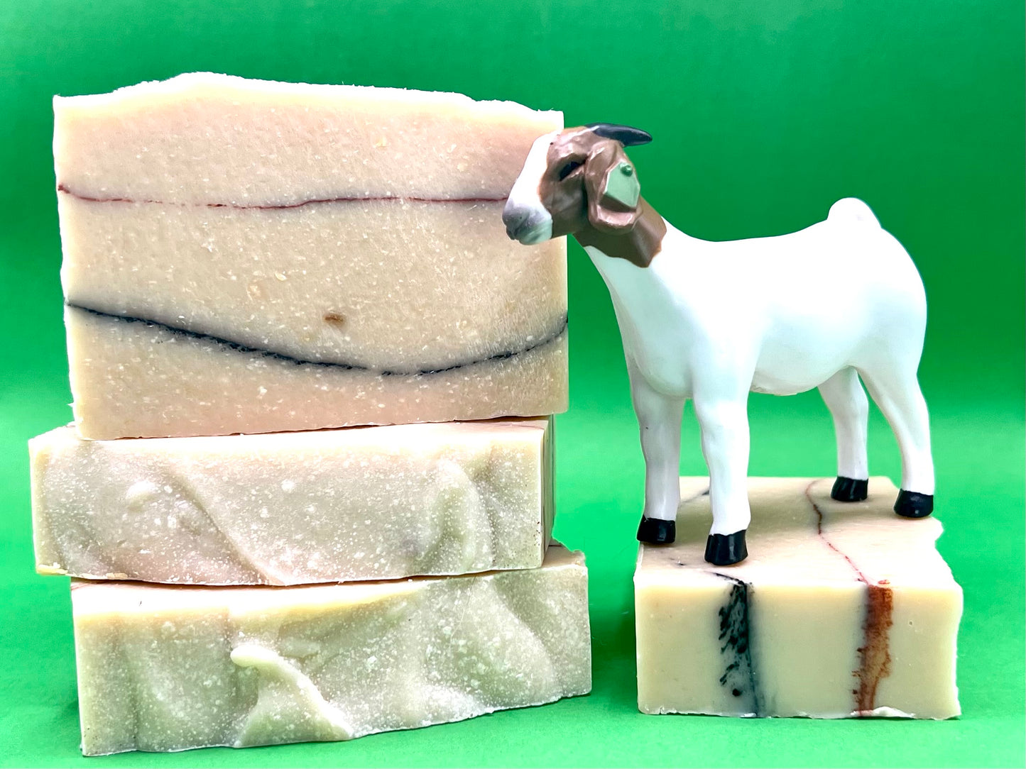 Four bars of goat milk soap. Three are stacked on the left side. The fourth is tipped on the right side with a plastic goat standing on top of it, facing left. The goat is white with a brown head. The soap is white with a red and blue stripe. The background is green.