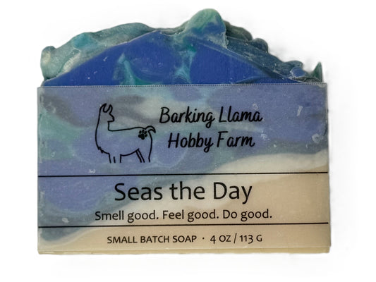 A small bar of handmade soap wrapped in a paper sleeve. The soap has a marbled blue and green top, and the label reads 'Barking Llama Hobby Farm' and 'Seas the Day'.