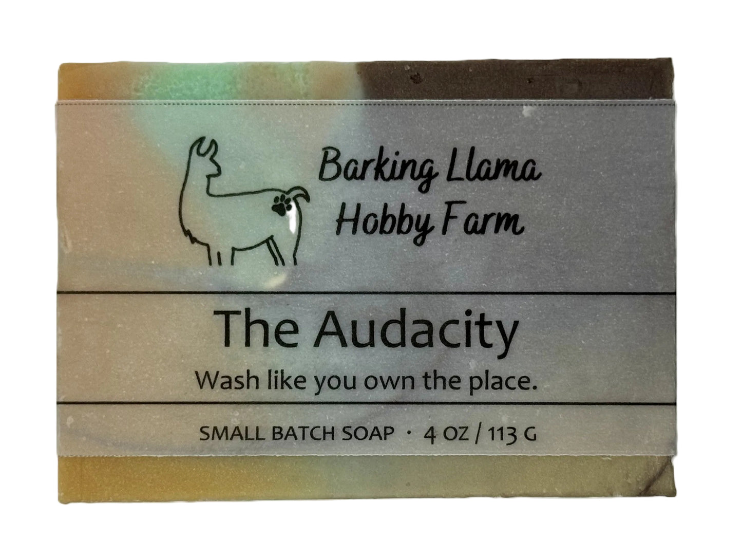 A rectangular bar of Barking Llama Hobby Farm 'The Audacity' small batch soap, featuring a silver-gray paper label with black text and a llama illustration, set against a plain white background. The soap is brown, orange, white, and turquoise swirled.