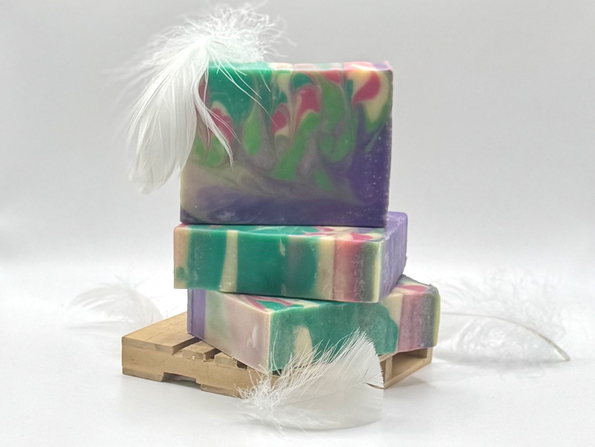 Three stacked handmade soap bars with a marbled swirl pattern of green, purple, pink, and cream, decorated with white feathers and displayed on a small wooden stand against a white background.