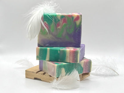 Three stacked handmade soap bars with a marbled swirl pattern of green, purple, pink, and cream, decorated with white feathers and displayed on a small wooden stand against a white background.