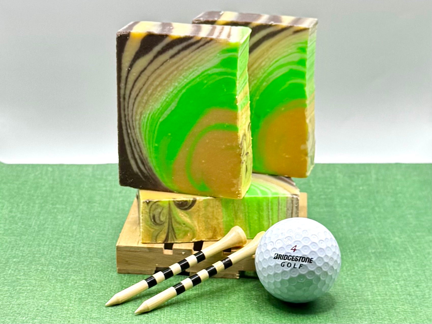 Two rectangular bars of marbled green and brown soap displayed on a small wooden stand, with two golf tees and a white Bridgestone golf ball on a green textured surface against a light gray background.