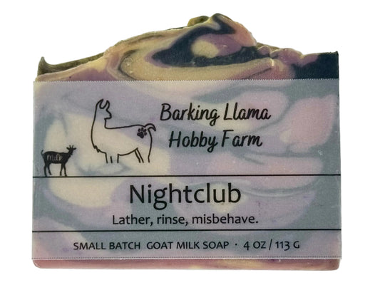 A bar of goat milk soap from Barking Llama Hobby Farm, labeled 'Nightclub'. The soap has a marbled pattern of cream, purple, and black, with a vellum paper label wrapped around it.