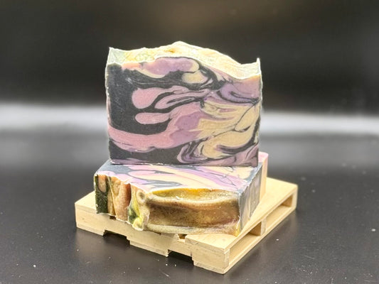 A handmade soap bar with a marbled pattern of black, purple, pink, and beige, displayed on a small wooden stand against a dark background.