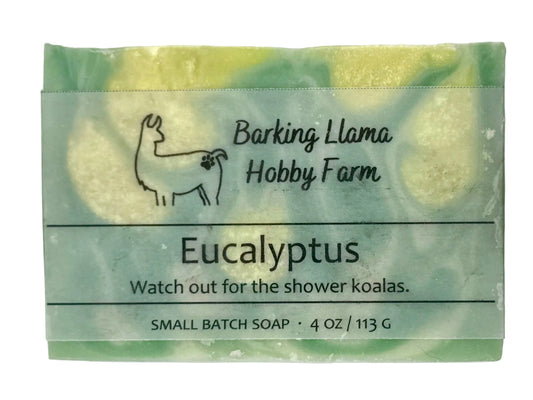 A small rectangular bar of Barking Llama Hobby Farm Eucalyptus soap with a clear paper label, showing a marbled green and pale yellow soap bar behind it.