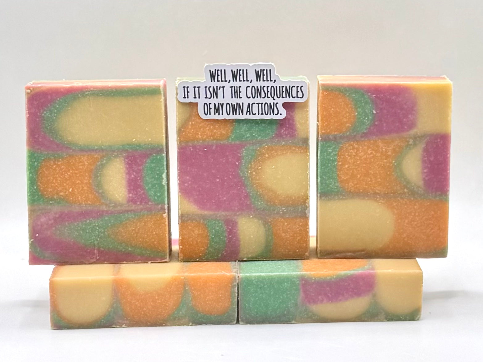 Five handmade rectangular bars of soap with a marbled swirl pattern of orange, purple, green, and cream, arranged on a white background. The center bar has a small white paper label with black text that reads, 'WELL, WELL, WELL, IF IT ISN'T THE CONSEQUENCES OF MY OWN ACTIONS.